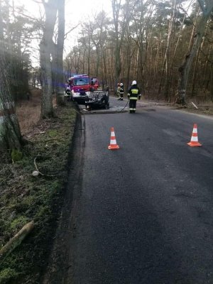 Na obrazie widać samochód, który dachował,  w wyniku uderzenia w drzewo. Na obrazie widoczni są też strażacy wykonujący czynności służbowe.