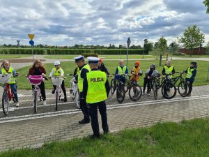 Na zdjęciu widać dzieci na rowerach, którzy zdawali egzamin na kartę rowerową. Tyłem odwróconych jest dwóch policjantów, którzy mają kamizelki odblaskowe z napisem Policja