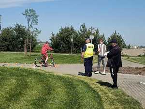 Na obrazie widać dziecko, które kieruje rowerem. Policjant stoi tyłem na zdjęciu, widoczna jest kamizelka odblaskowa z napisem Policja