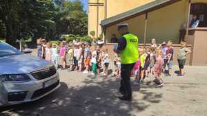 Na zdjęciu widać policjanta, w kamizelce z napisem POLICJA. Dzieci odwrócone są przodem do fotografa, policjant tyłem