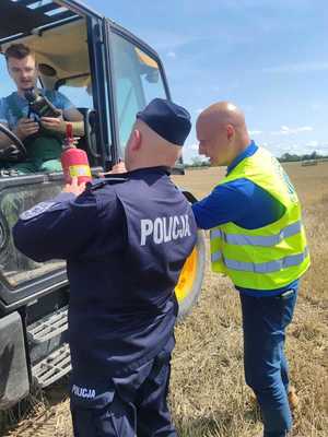 Na zdjęciu widać policjanta, kontrolującego gaśnicę w maszynie rolniczej. Na zdjęciu obok policjanta jest przedstawiciel PIP-u. Ubrany w odblaskową kamizelkę. Widać też maszynę rolniczą, a w niej siedzącego operatora.