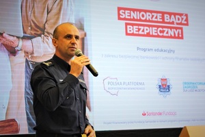 Na zdjęciu widać policjanta, trzymającego mikrofon. W tle napis: Seniorze bądź bezpieczny&quot;