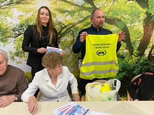 Na obrazie widać policjanta, który trzyma kamizelkę odblaskową z napisem WORD Kalisz. Odbyło się spotkanie z seniorami na temat bezpieczeństwa. Na zdjęciu widać seniorów, którzy siedzą na krzesłach  i osoby, które opiekują się seniorami.