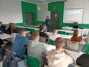 Na zdjęciu widać uczniów klas 4 z ZST w Pleszewie podczas spotkania, dotyczącego promocji zawodu policjanta. Uczniowie siedzą w ławkach tyłem do zdjęcia. Na obrazie widać policjantkę, która prowadzi tą prelekcję. Za jej plecami znajduje się tablica, na której został wyświetlony plakat &quot;Zostań jednym z nas&quot;. Na plakacie widać czterech policjantów, ustawionych obok siebie.