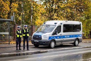 Na obrazie widać radiowóz, a na chodniku dwóch policjantów, którzy stoją przodem do zdjęcia