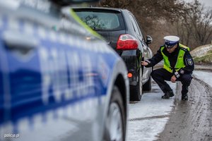 Na obrazie widać samochód, policjant sprawdza opony. Za samochodem stoi radiowóz