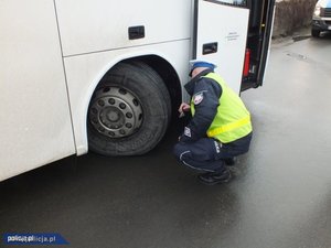 Na obrazie widać autobus koloru białego, obok policjant, który sprawdza ogumienie