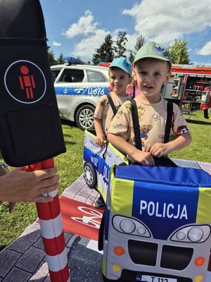 Na zdjęciu widać dwóch chłopców którzy są w autochodziku policyjnym. Uśmiechają się do zdjęcia. Widać też sygnalizację świetlną ze światłem czerwonym.