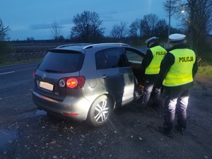 Na obrazie widać dwóch policjantów, którzy stoją tyłem do zdjęcia. Przed nimi stoi samochód Volkswagen. Policjanci są ubrani w kamizelki odblaskowe.