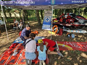 Na obrazie widać policjanta, który klęczy przy fantomie i wspólnie z ratownikami medycznymi prezentuje uczestnikom festynu jak należy wykonywać resuscytacje krążeniowo oddechową