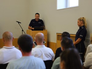 Na zdjęciu widać policjantów w mundurach i w ubraniach cywilnych. Policjanci w mundurach stoją, a w ubraniach cywilnych siedzą tyłem do zdjęcia.