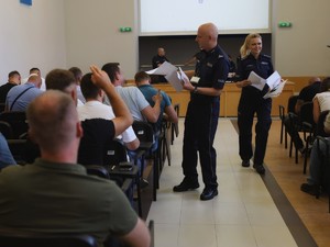 Na sali rozdaje policjant w mundurze testy. Policjanci bez munduru siedzą tyłem do zdjęcia na krzesłach