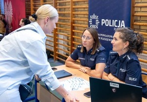 Na zdjęciu widać dwie policjantki, którzy siedzą przy stoliku, a przed nimi stoi kobieta.