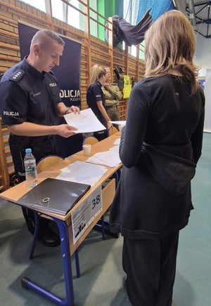 Na zdjęciu widać policjanta za stolikiem. Tyłem do zdjęcia stoi dziewczyna, która rozmawia z policjantem na temat rekrutacji do szkoły.