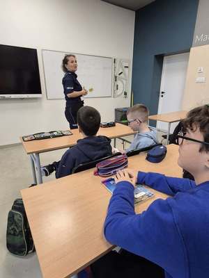 Na obrazie widać policjantkę i uczniów. Policjantka stoi, a uczniowie siedzą na krzesłach. Zdjęcie wykonane zostało podczas prowadzonej prelekcji. Uczniowie skierowani są tyłem do zdjęcia, a policjantka stoi bokiem do zdjęcia.