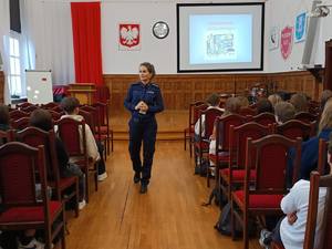 Na obrazie widać policjantkę, stojącą i prowadzącą prelekcję na temat uzależnień. Na krzesłach siedzą uczniowie tyłem do zdjęcia.