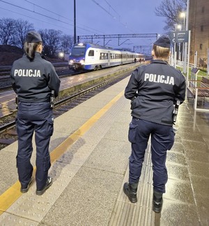 Na obrazie widać policjantki na dworcu kolejowym w Pleszewie. Idą wzdłuż torów i patrolują teren.. W oddali pociąg. Funkcjonariuszki są skierowane tyłem do zdjęcia