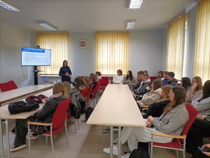 Na obrazie widać policjantkę, która prowadzi spotkanie dotyczące promocji zawodu policjanta. Spotkanie odbywa się na sali. W ławkach siedzą uczniowie ZST w Pleszewie.