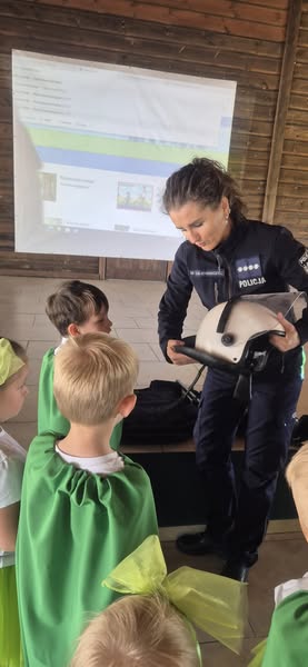 Na obrazie widać policjantkę, która prezentuje sprzęt używany do codziennej służby. Jest to kask, wykorzystywany podczas meczów.