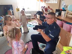 Na obrazie widać policjanta, który pokazuje dzieciom czapki policyjne, policjant siedzi na krześle, a wokół niego są dzieci