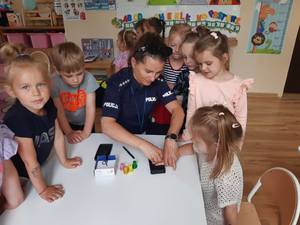 Na obrazie widać policjantkę, która pokazuje dzieciom jak należy pobierać odciski palców.