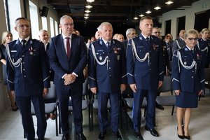W tle widać policjantów, którzy stoją obok siebie.