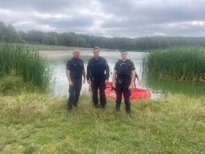 Na obrazie widać policjantów umundurowanych i strażaka, a w tle zalew gołuchowski i łódź motorowa.
