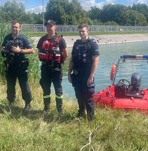 Na obrazie widać policjantów umundurowanych i strażaka, a w tle zalew gołuchowski i łódź motorowa.