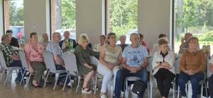 Na obrazie widać seniorów którzy siedzą na krzesłach, przy stołach. Są ustawieni przodem do zdjęcia.