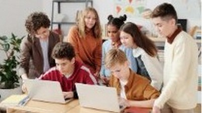 Na obrazie widać kilka osób, które siedzą lub stoją przy biurku, na którym znajduje się laptop