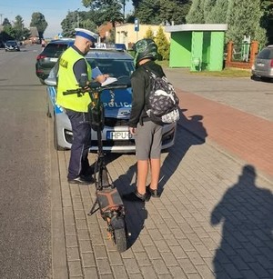 Na obrazie widać policjanta, radiowóz policyjny i chłopca na hulajnodze, Chłopiec ubrany ma na głowie kask.