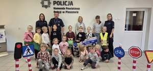 Na zdjęciu widać policjantów, dzieci z przedszkola i opiekunów. Na zdjęciu widoczne są znaki drogowe, autochodziki, a za plecami na ścianie napis Komenda Powiatowa Policji w Pleszewie.
