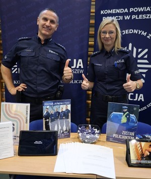 Policjanci stoją na tle banerów Komenda Powiatowa Policji w Pleszewie i Zostań jednym z nas. Jest policjant i policjantka. Zdjęcie wykonane podczas targów pracy.