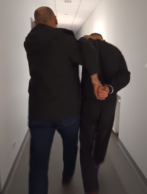 Na obrazie widać mężczyznę i policjanta który prowadzi zatrzymanego. W tle widać korytarz.