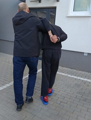 Na obrazie widać mężczyznę i policjanta który prowadzi zatrzymanego. W tle widać drzwi.