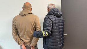 Widać policjanta ubranego w cywilną kurtkę z opaską na rękawie. Trzyma on zatrzymanego pod rękę. Zatrzymany ma kajdanki na dłoniach z tyłu.