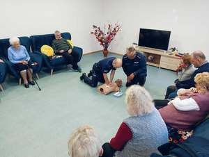 Policjanci klęczą przy manekinie i wykonują resuscytację krążeniowo-oddechową. wokół są skupieni seniorzy, którzy siedzą na krzesełkach.