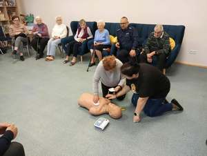 Seniorzy klęczą przy manekinie i wykonują resuscytację krążeniowo-oddechową. Wokół są skupieni seniorzy, którzy siedzą na krzesełkach.