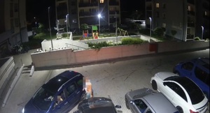 Na obrazie widać pojazdy i mężczyznę, który przechodzi obok innego pojazdu i rysuje maskę pojazdu