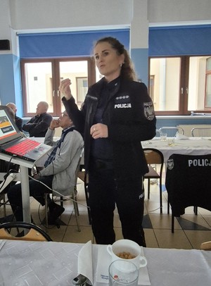 Policjantka prowadzi prelekcję na temat bezpieczeństwa osób starszych. Seniorzy siedzą przy stole. Policjantka skierowana jest przodem do zdjęcia.