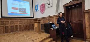 Na obrazie widać policjantkę, która prowadzi zajęcia z rodzicami o bezpieczeństwie w ruchu drogowym. Policjantka stoi, a za nią znajduje się komputer i wyświetlana jest prezentacja na temat przyczyn i skutków wypadków drogowych.