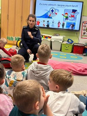 Na zdjęciu policjantka, która znajduje się przed dziećmi z przedszkola. Za policjantką znajduje się tablica, na której wyświetlany jest napis "Życzymy bezpiecznych ferii". Z przodu zdjęcia, tyłem znajdują się dzieci, które siedzą na dywanie.