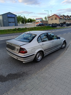 Na obrazie widać samochód marki BMW koloru jasnego, który ma łyse opony i uszkodzone reflektory. Samochód stoi na drodze.